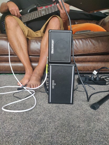 Two guitar amplifiers (one branded 'Blackstar', the other 'BOSS') stacked on to of one another on carpet in front of a leather couch. The legs and bass of our bass player is showing next to/behind the 'stack' for scale (the amps go part way up his shin, height-wise). This was our Spinal Tap moment. *Stonehenge*!