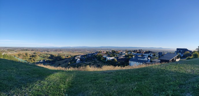 View from Westmoreland hill looking west-north-west towards the Southern Alps across the Canterbury plains. The sky is cloudless blue in the morning light. The foreground grassy hillside is a lush green.