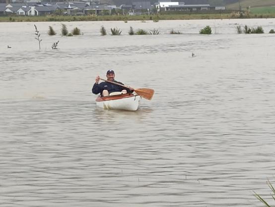 Rainy photo of a bloke paddling an orange and white canoe with a single paddle on a lake with reeds poking out. He's paddling towards the camera, about 15m away.