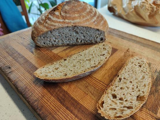 Two slices of a round unbleached flour sourdough loaf showing the crust and crumb. On a wood cutting board on a stone benchtop. Ingredients: locally ground unbleached wheat flour, rye sourdough starter, salt, and a bit of plain greek yoghurt, water.